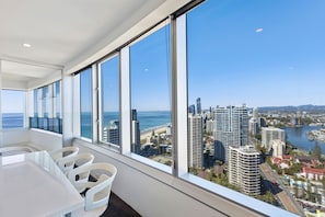 Une chambre d’hôtel moderne dotée de grandes fenêtres donnant sur la ville, d’un coin repas avec des chaises blanches et d’une vue sur l’océan.