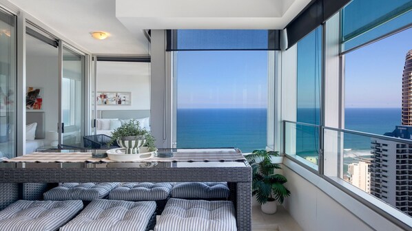A modern interior with a large window overlooking the ocean, a seating area with cushions, and a kitchen with a view.