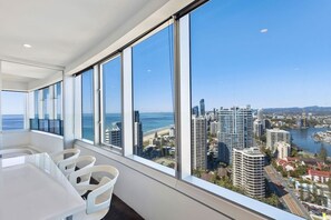 A modern hotel room with large windows, a dining area, and a view of a cityscape and coastline. - Luxury 3 Bedroom Apartment with Ocean Views (Surfers Paradise)