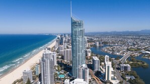 A high-rise building with a unique curved design, surrounded by a coastal cityscape with a beach and a river.