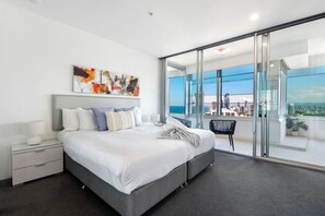A modern hotel room with a large bed, a nightstand, a chair, and a balcony with a city view. - Luxury 3 Bedroom Apartment with Ocean Views (Surfers Paradise)