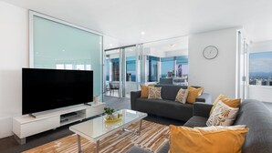 A modern living room with a flat-screen TV, a sofa set, a glass coffee table, and a city view through large windows.