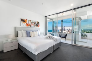 A modern bedroom with a large bed, a bedside table, a chair, and a view of the city through floor-to-ceiling windows. - Luxury 3 Bedroom Apartment with Ocean Views (Surfers Paradise)