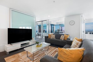 Un salon moderne doté d’un téléviseur à écran plat, d’une table basse en verre, d’un canapé gris agrémenté de coussins à motifs et offrant une vue sur la ville grâce à de grandes fenêtres.
