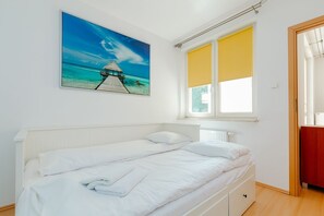 A bedroom with a bed, a window with yellow blinds, a white radiator, and a painting of a beach scene. - ShortStayPoland Św. Wincentego B7 (Warsaw)