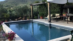 A rectangular swimming pool with a pergola and outdoor seating area, surrounded by greenery and a mountainous backdrop.
