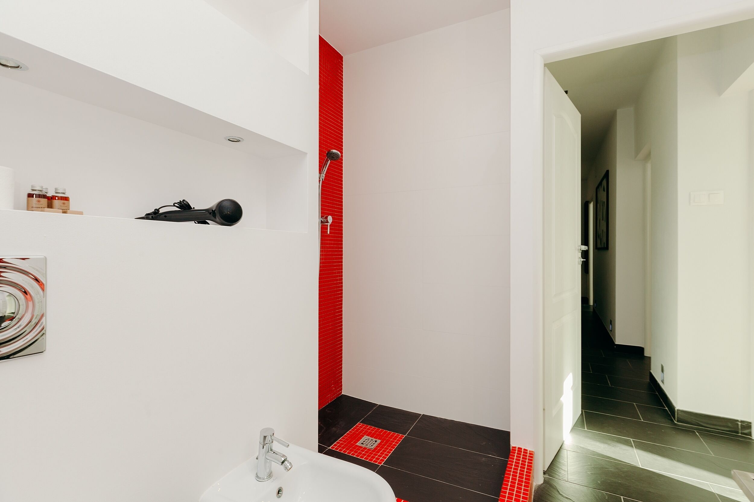 Une salle de bain moderne avec un lavabo blanc, une douche aux carreaux rouges et un sèche-cheveux noir posé sur une étagère.