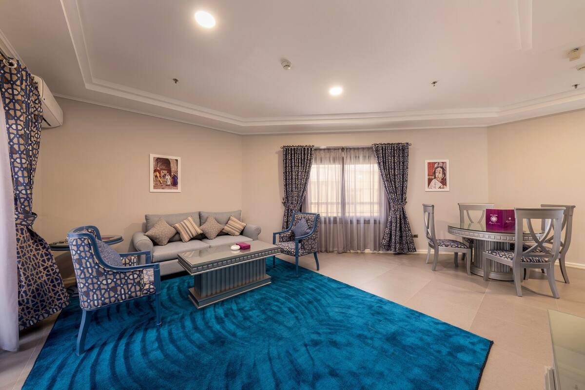 Un salon avec un tapis bleu, un canapé gris, des fauteuils à motifs, une table basse en verre et un coin repas comprenant une table et des chaises.