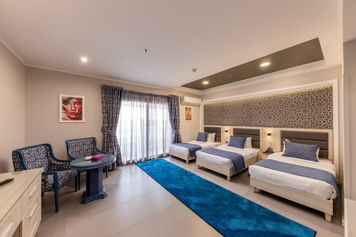 Une chambre d’hôtel avec deux lits simples, un bureau avec une chaise, une petite table et une moquette bleue.