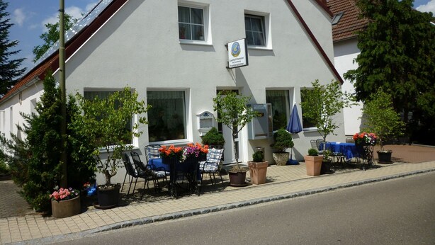 A two-story building with a sign indicating it is a pension. There are outdoor seating areas with tables and chairs, and potted plants around. - Gasthaus - Pension Am Buchberg (Mönchsdeggingen)