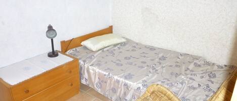 A small, neatly arranged bedroom with a single bed, a wooden nightstand, a wicker chair, and a desk with a lamp.