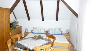 A cozy bedroom with a sloped ceiling, wooden beams, a bed with striped bedding, a bedside table with lamps, a wooden dresser, and a small table with a glass top. - Gasthaus - Pension Am Buchberg (Mönchsdeggingen)