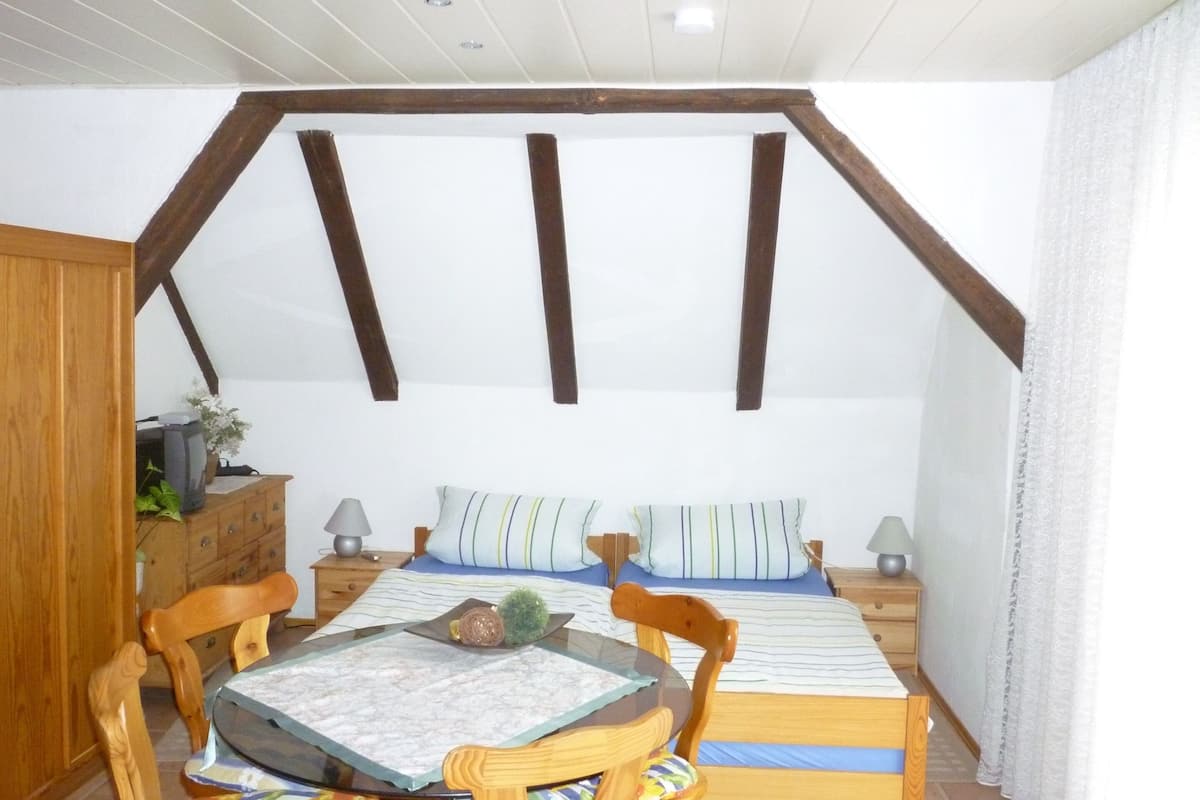 A cozy bedroom with a sloped ceiling, wooden beams, a bed with striped bedding, a bedside table with lamps, a wooden dresser, and a small table with a glass top.