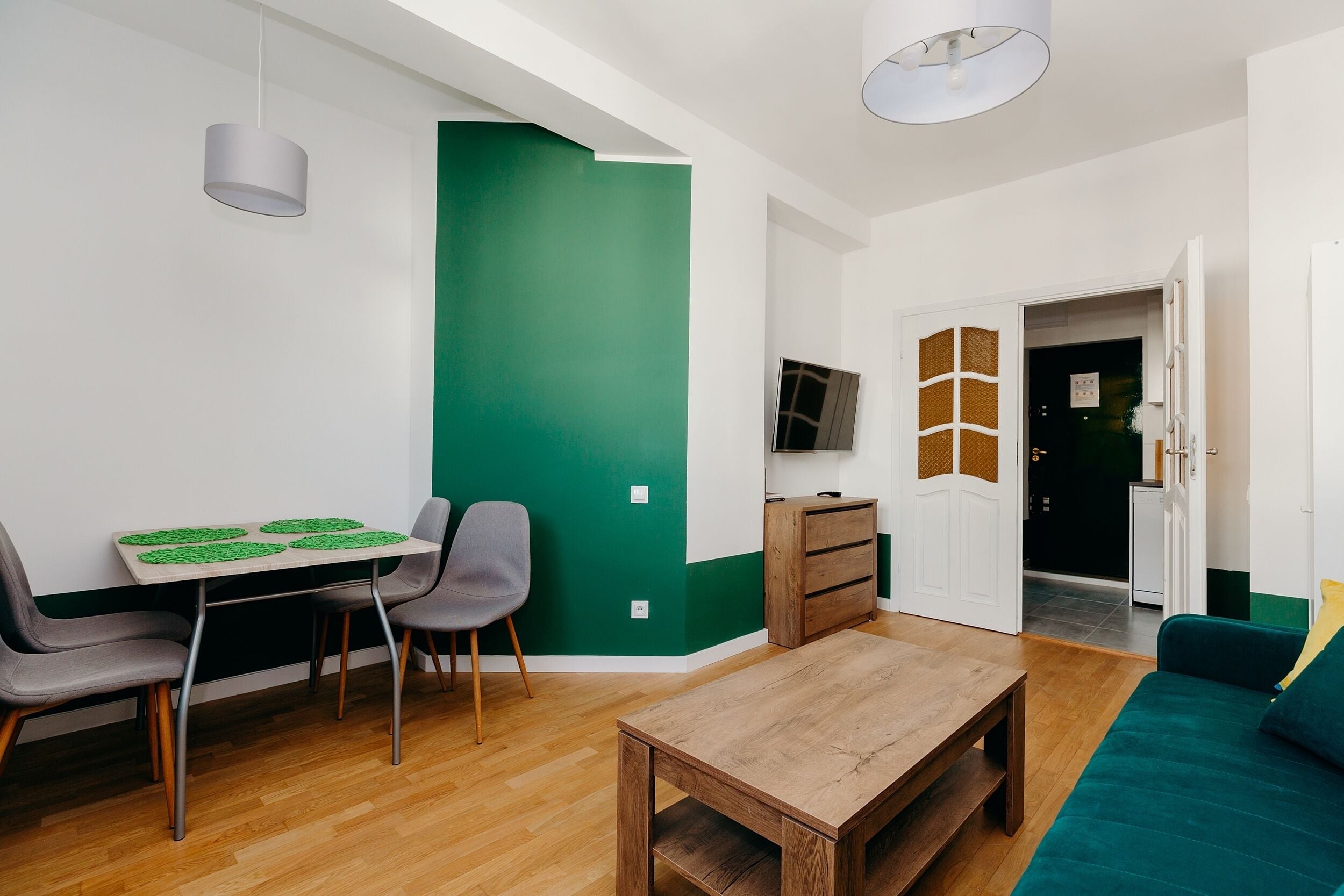 A modern living room with a wooden table, two grey chairs, a green accent wall, and a teal sofa.