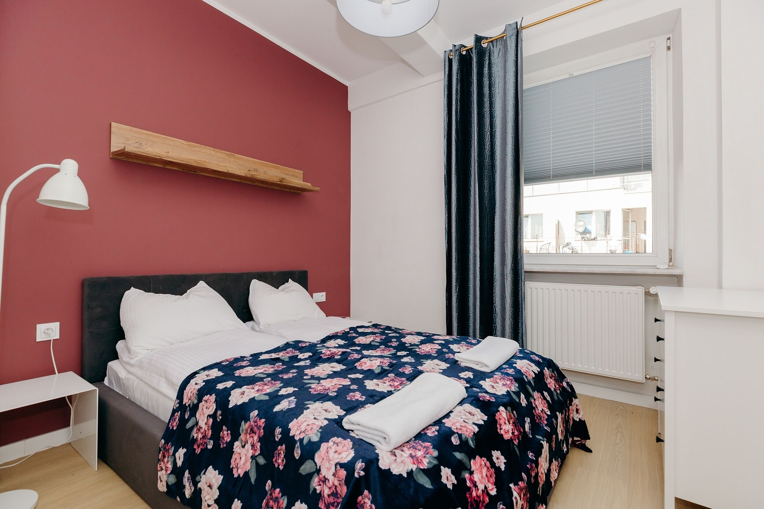 Une chambre à coucher avec un lit, une étagère en bois, un bureau blanc, une fenêtre avec des rideaux et un radiateur.