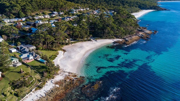 An aerial view of a coastal area with a sandy beach, clear blue waters, and a residential area with houses. - The Green Whaler, Hyams Beach (Hyams Beach)