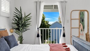 A bedroom with a bed, a large potted plant, a mirror, and a view of a blue house through a window with white curtains. - The Green Whaler, Hyams Beach (Hyams Beach)