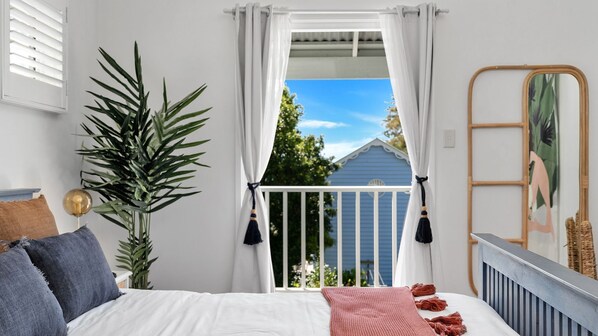 A bedroom with a bed, a large potted plant, a mirror, and a view of a blue house through a window with white curtains.