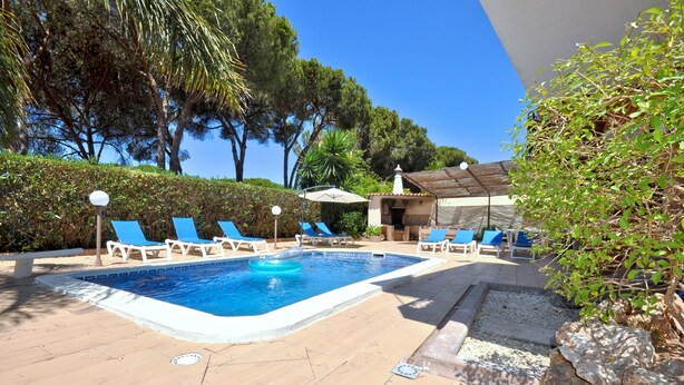 Une piscine entourée d’arbres et d’un bâtiment, avec des chaises longues et des parasols.