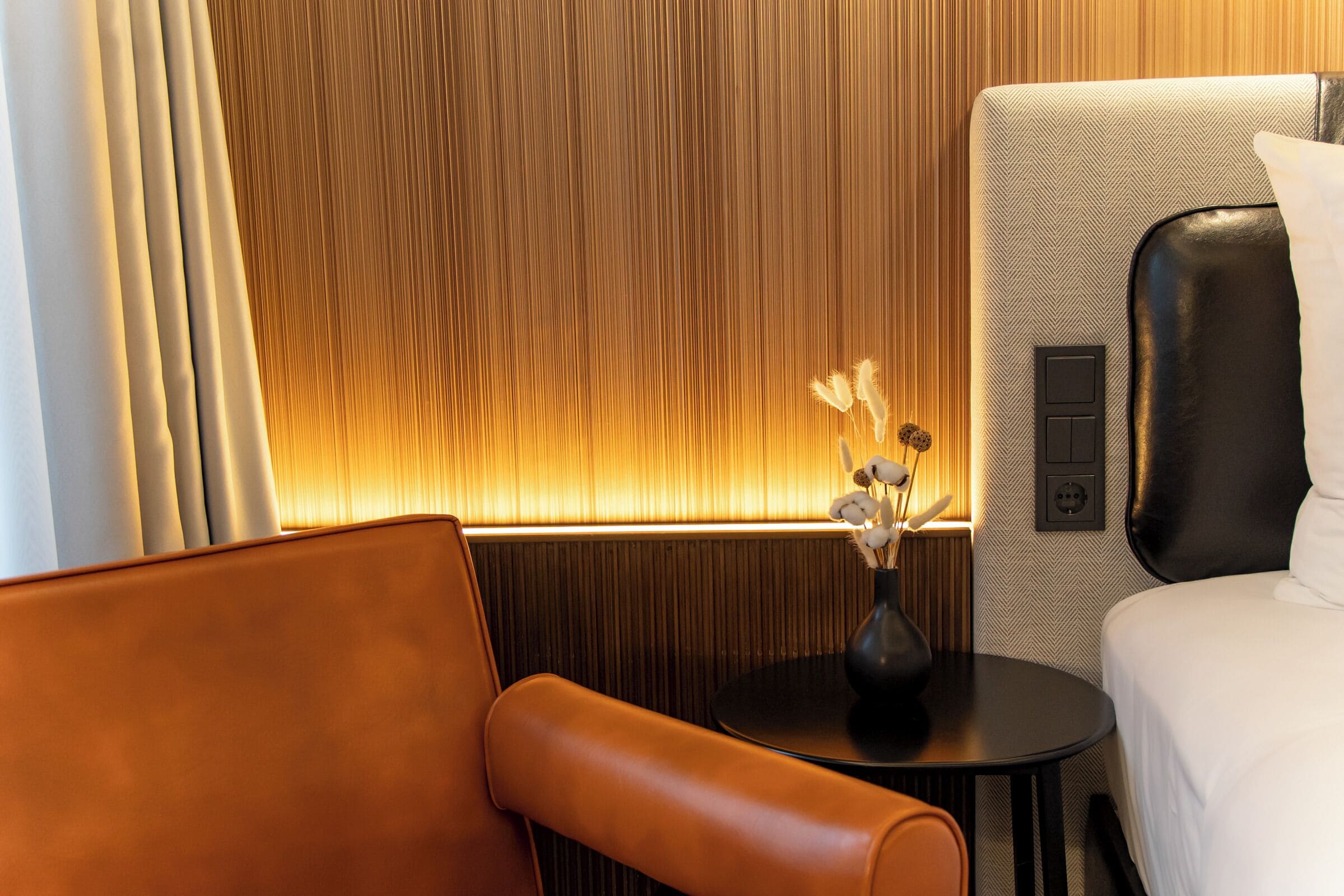 Una habitación de hotel moderna con una cama grande, un sillón de cuero, una mesita redonda con un jarrón de flores secas y un panel de pared con textura.
