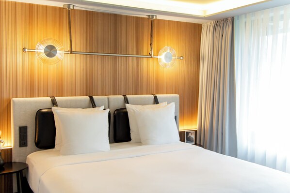Una habitación de hotel con una cama grande, un cabecero con una lámpara, una mesita de noche y una ventana con cortinas.