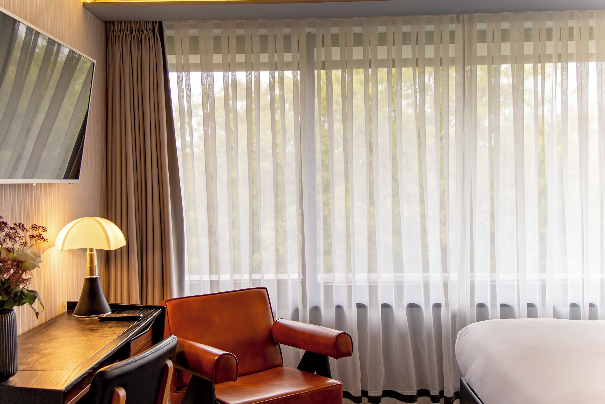 A hotel room with a bed, a desk with a lamp, a chair, and sheer curtains.