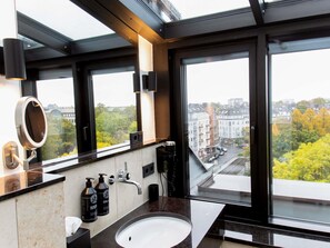 Un baño moderno con un ventanal grande, un espejo redondo, un lavamanos y dos botellas de champú.
