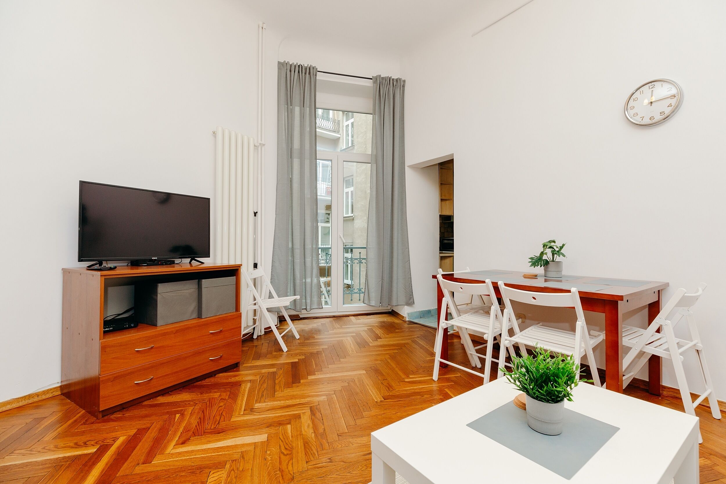 A modern room with a wooden herringbone floor, a TV on a wooden cabinet, white chairs, a dining table, and a clock on the wall.