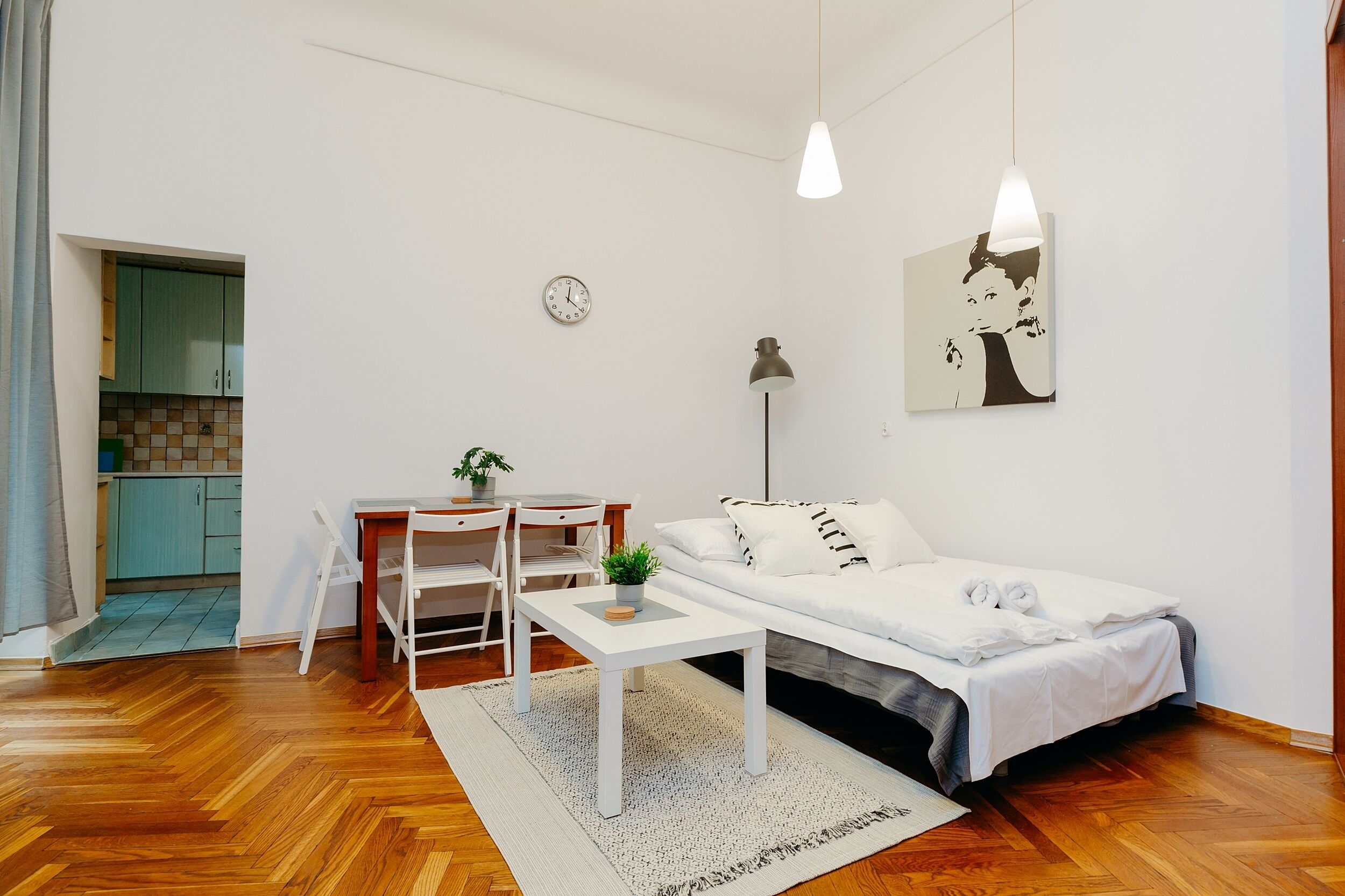 Une pièce de taille modeste comprenant un lit, un bureau, une chaise, une petite table avec une plante, une horloge et une œuvre d’art murale.