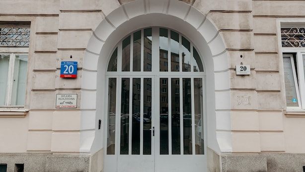 Entrada de un edificio con un amplio arco, letreros con números y un acceso de vidrio con paneles verticales.