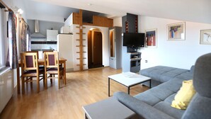 Una sala moderna con un sofá gris, una mesa de comedor de madera con sillas, un televisor y una cocina al fondo.