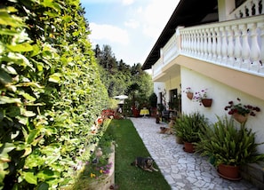A garden pathway leading to a building with a white exterior and a balcony. There are potted plants and a dog lying on the grass.