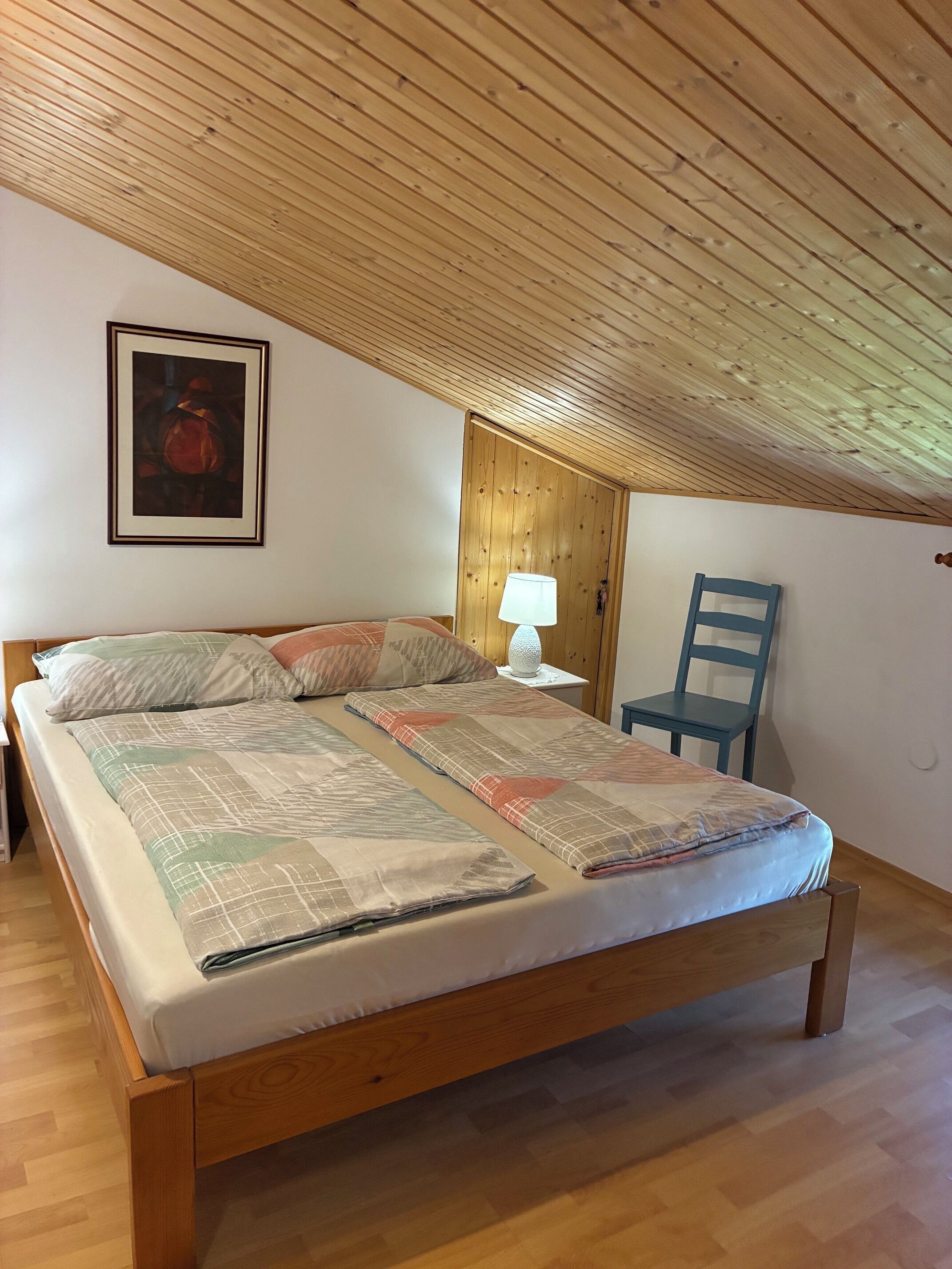 Un dormitorio con techo de madera, una cama con cubrecama estampado, una silla y una mesita de noche con lámpara.