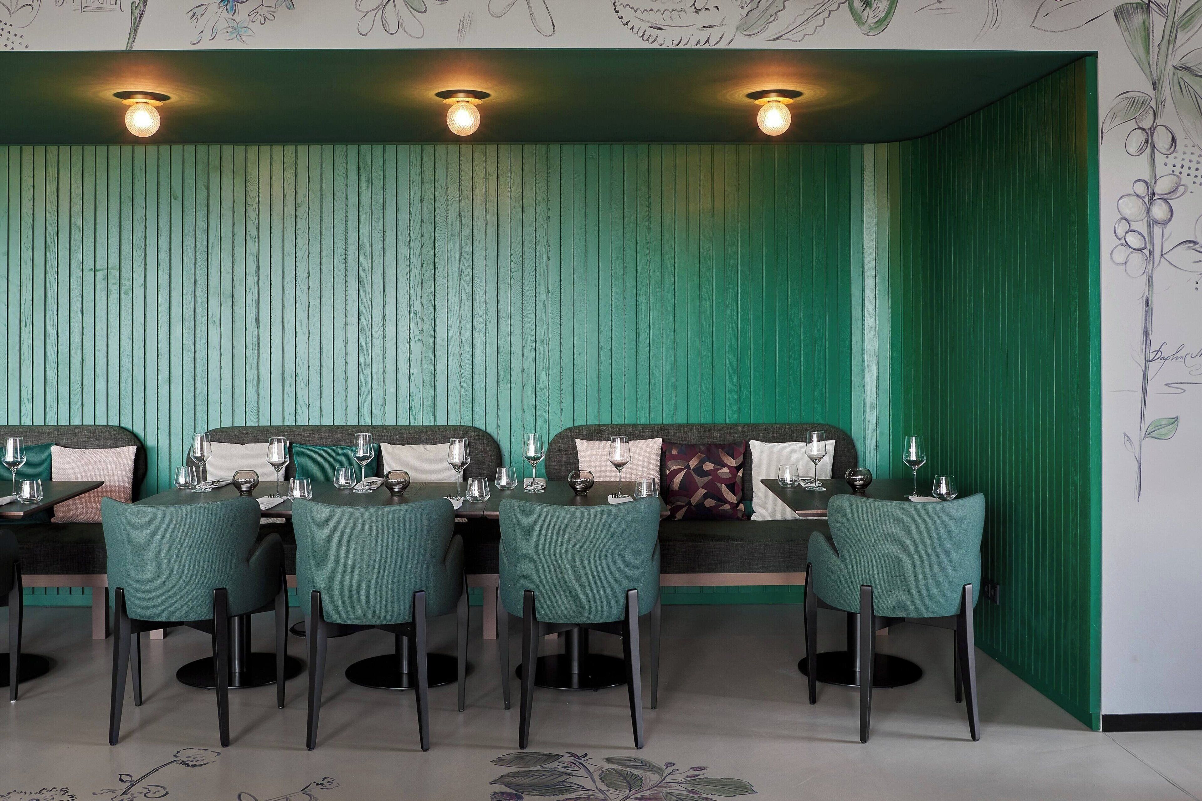 A restaurant with a green-themed interior, featuring a long dining table with chairs, a sofa, and a wall with a botanical mural.