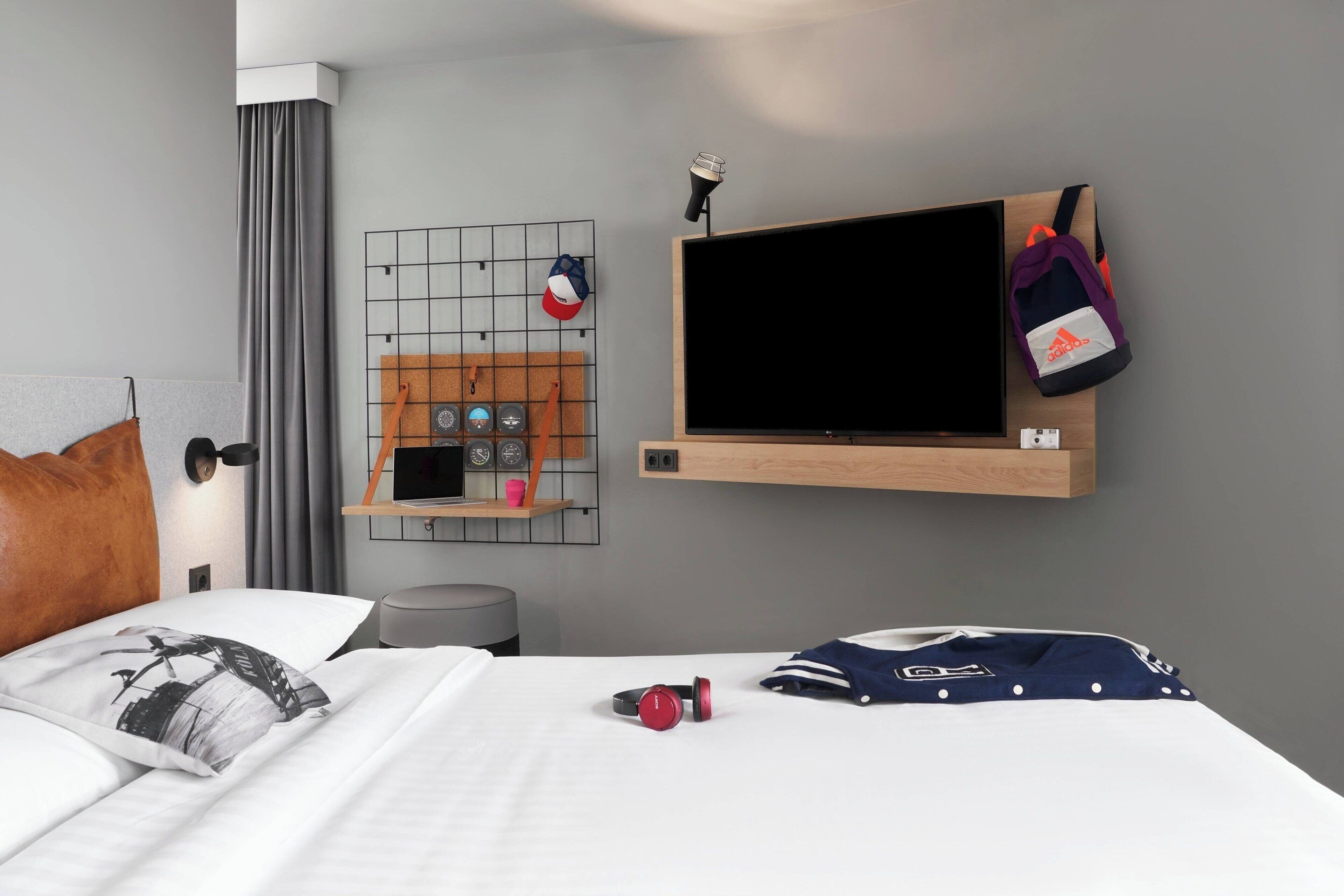 A modern hotel room with a bed, a mounted TV, a wall-mounted shelf with a hat and sunglasses, and a small desk with a lamp.