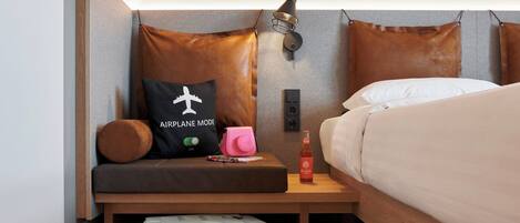 Une chambre d’hôtel de taille réduite, comprenant un lit, une table de chevet, un petit coin salon avec un coussin, une lampe et une valise.