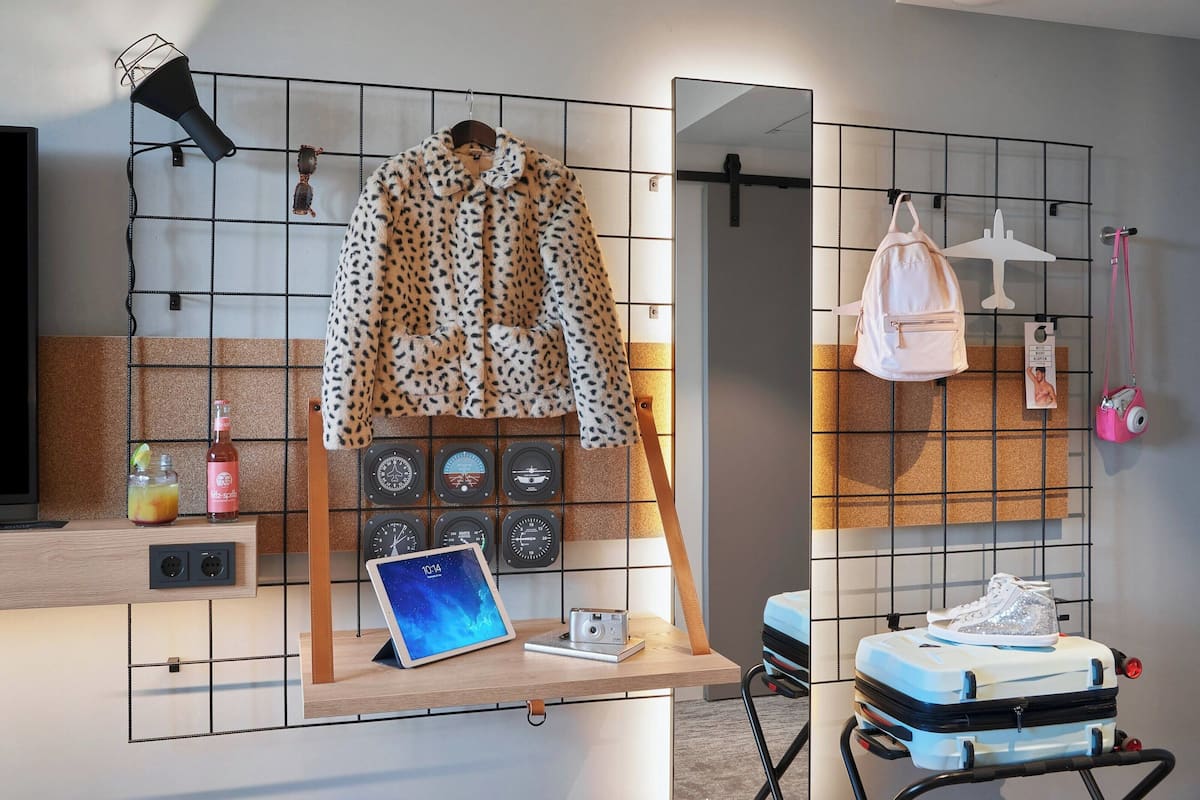 A room with a wooden desk, a coat rack, a suitcase, and a wall with clocks and a mirror.