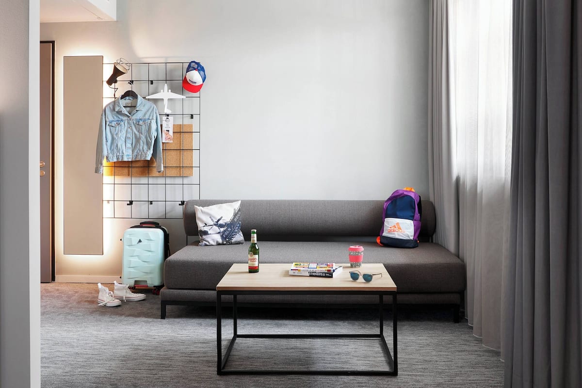 A modern hotel room with a grey sofa, a wooden coffee table, a suitcase, a backpack, and a wall-mounted rack with clothes and a hat.