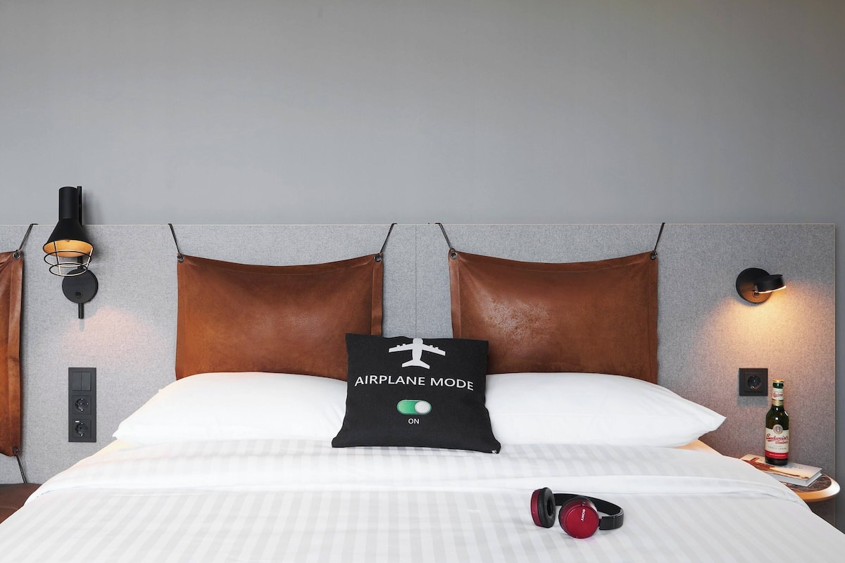 A hotel room with a bed, a pillow with "AIRPLANE MODE" text, a bottle of beer, and headphones on the bed.