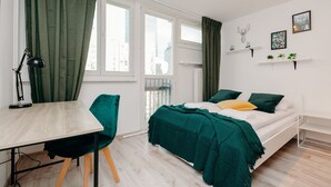 A modern bedroom with a bed, a desk, and a chair. There are windows with curtains, a wall-mounted shelf with plants, and a picture. - ShortStayPoland Krochmalna B17 (Warsaw)