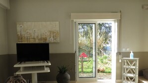 Un salon avec un téléviseur à écran plat posé sur un meuble blanc, une plante en pot, une lanterne décorative et une porte donnant sur un espace extérieur agrémenté d’un parasol rouge.