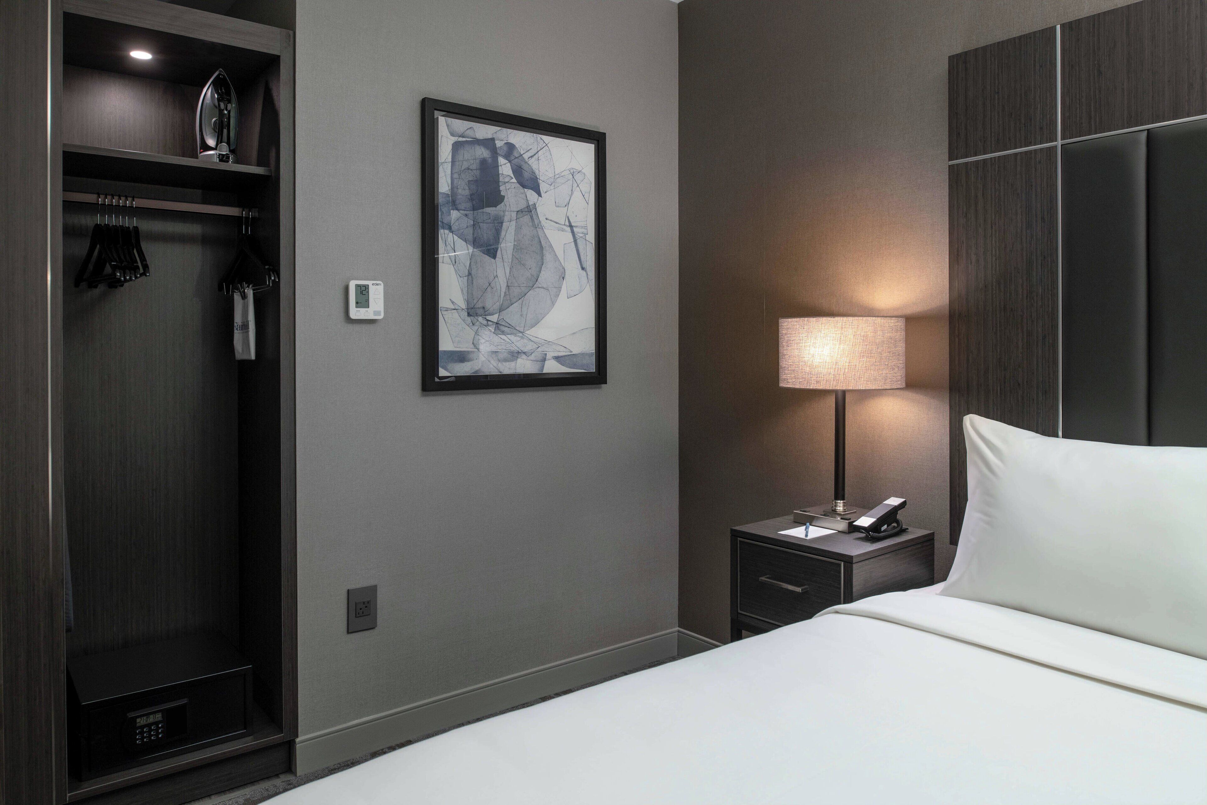 A modern hotel room with a bed, a nightstand with a lamp, a wardrobe, and a framed abstract artwork on the wall.