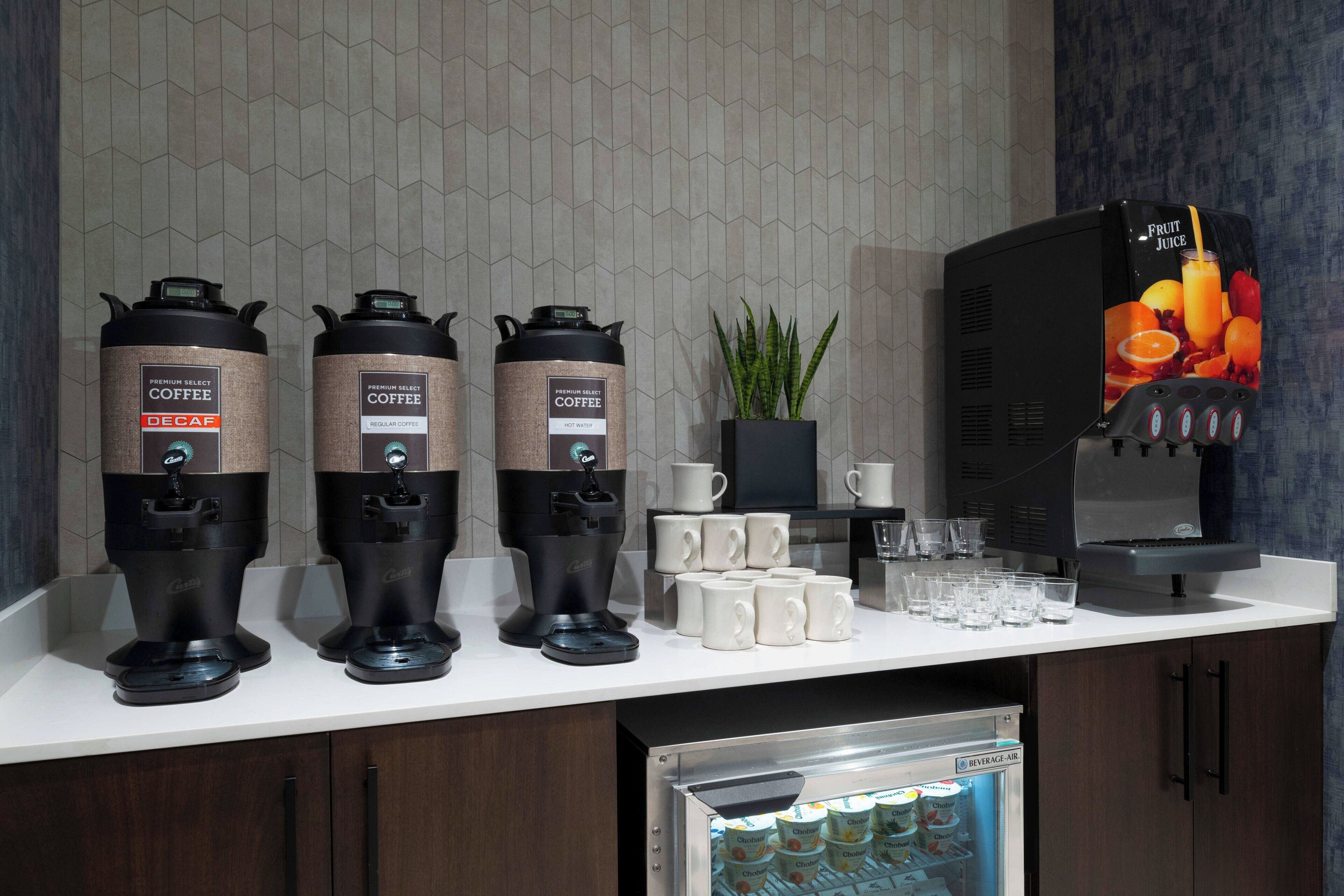 A coffee station with three coffee dispensers, a refrigerator with bottled water, and a fruit juice dispenser.