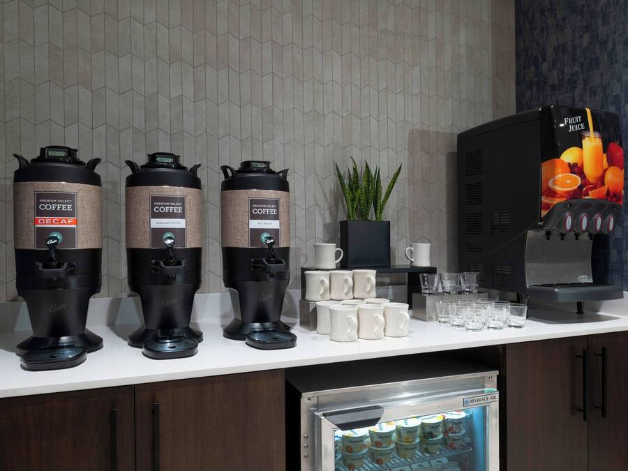 A coffee station with three coffee dispensers, a refrigerator with bottled water, and a fruit juice dispenser.