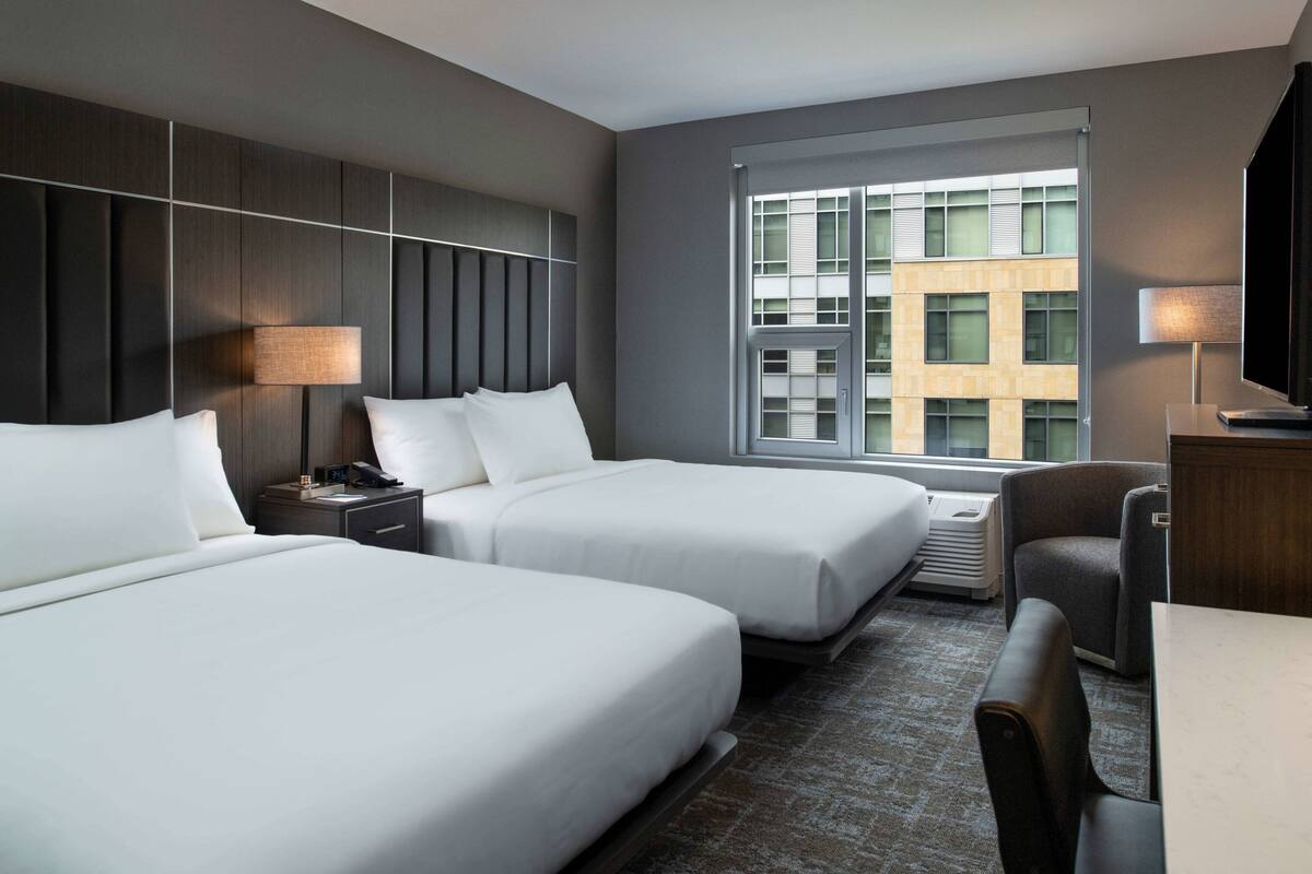 A hotel room with two beds, a desk, a chair, a TV, and a window with a view of a building.