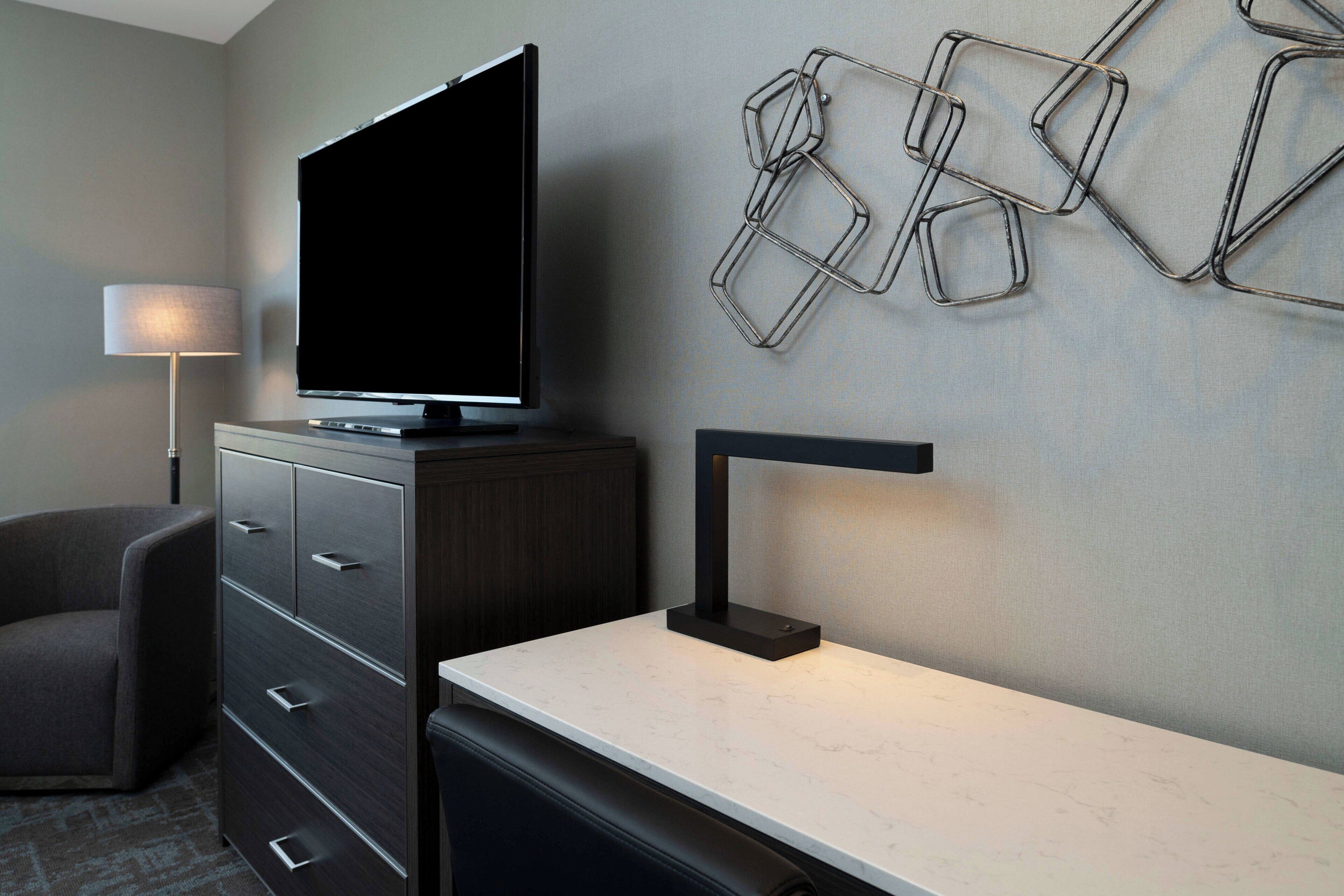 A modern hotel room with a flat-screen TV on a cabinet, a desk with a lamp, a chair, and a wall-mounted geometric light fixture.