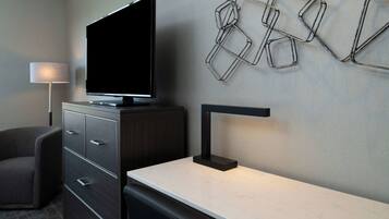 A modern hotel room with a flat-screen TV on a cabinet, a desk with a lamp, a chair, and a wall-mounted geometric light fixture.