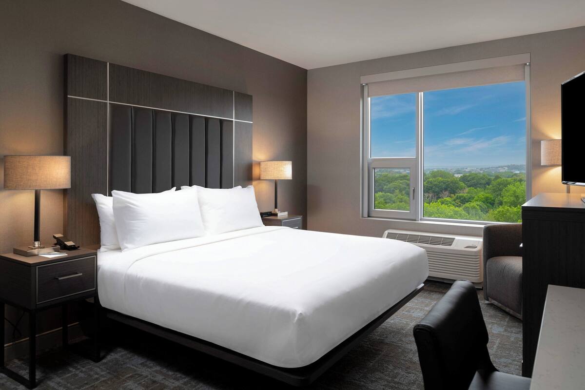 A modern hotel room with a large bed, two bedside tables with lamps, a desk with a chair, and a large window offering a view of greenery.