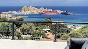Eine Küstenstadt mit einem markanten roten Schiff namens 'Corsica tim.ea', das in der Nähe einer felsigen Insel vor Anker liegt.