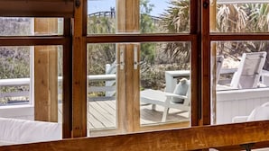 A wooden bench with white cushions and a view of outdoor furniture through large windows. - One-of-a-Kind Trolley Home, Oceanfront w/ Dune View by Tybee Cottages (Tybee Island)